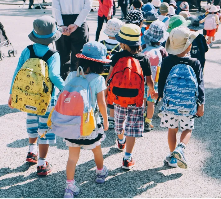 Group of young children walking together. They wear colorful backpacks and hats. 