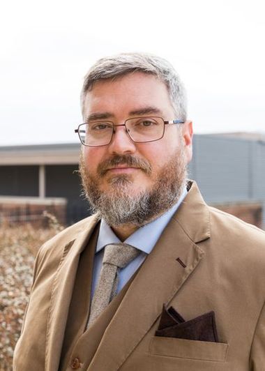 Male professor with greying beard and hair wears wire rimmed glasses, a brown sports coat, vest and tie. 